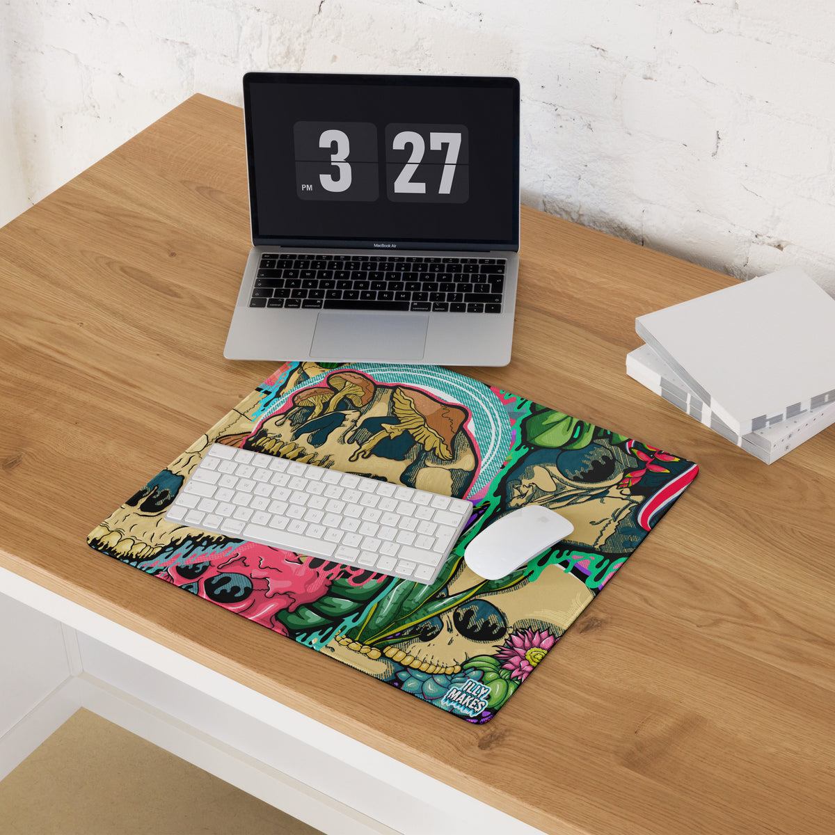 Product mockup of Illymakes gaming mousepad with a colorful collage of dripping skulls, mushrooms, and florals, shown with a keyboard, mouse, and headphones.